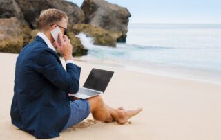 A professional in a suit using a laptop and phone on the beach.